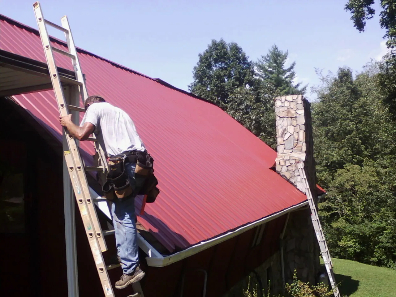 Roofer on a ladder beside a red metal roof and stone chimney — Soffit & Fascia Repair by Roofing Brothers in Nassau